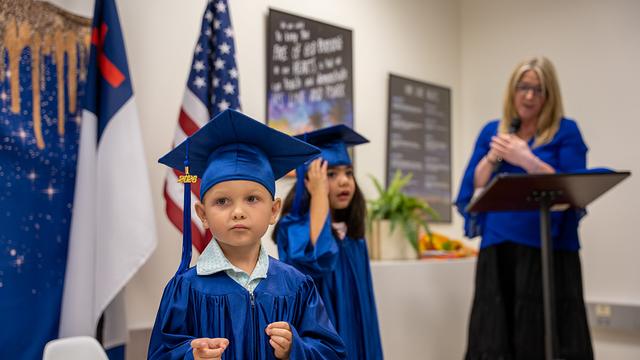 A graduate in cap and gown at the front of the room beside the flags
