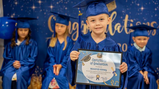 A graduate proudly holding up a certificate while classmates sit behind
