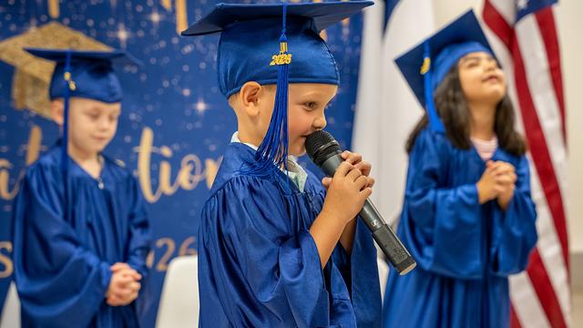A graduate in cap and gown speaking into a microphone while classmates look on