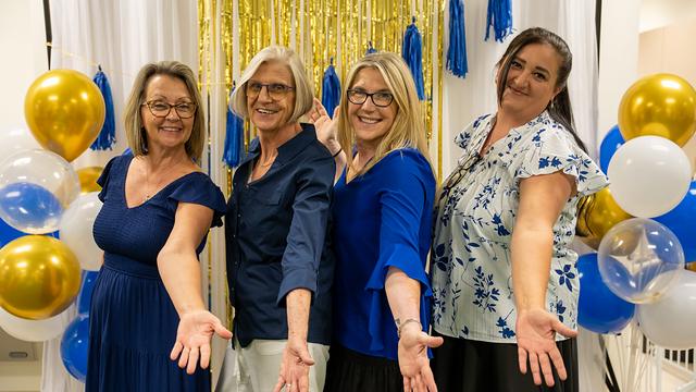 Four women in blue posing with open arms beside blue and gold balloons