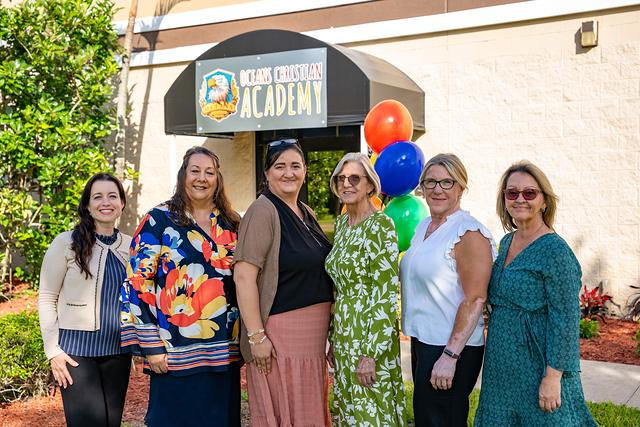 Six women smiling together outside the school entrance beside the balloons