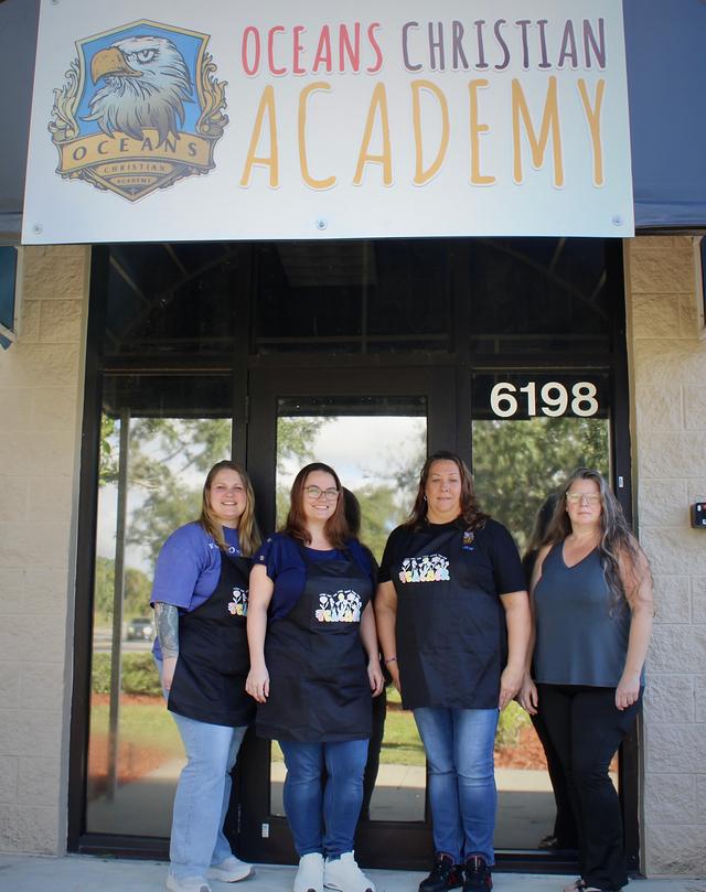 Four women, three in black teacher aprons, at the front door under the Oceans Christian Academy sign