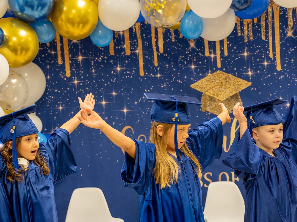 OCA preschool graduates in blue caps and gowns cheering in front of blue and gold balloons