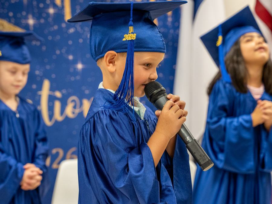 A preschool graduate in a blue cap and gown speaking into a microphone at graduation